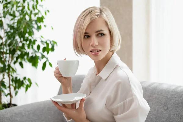 Junge schöne blonde Mädchen mit kurzen Haaren in einem weißen Hemd und Brille sitzt auf dem Sofa in hell im Büro gegen das Fenster. hält eine weiße Tasse und Untertasse. Kaffee oder Tee in einem — Stockfoto