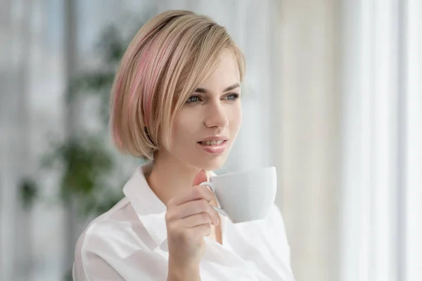 Молодая красивая блондинка с короткими волосами в белой рубашке и очках сидит на диване в ярком офисе у окна. Держит белую чашку и блюдце. Пить кофе или чай в — стоковое фото
