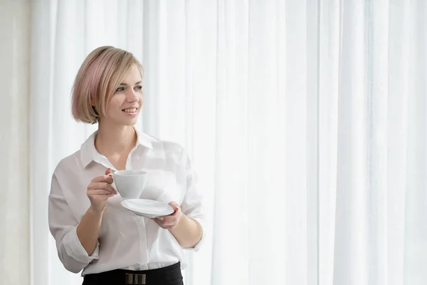 Junge schöne blonde Mädchen mit kurzen Haaren in einem weißen Hemd und Brille sitzt auf dem Sofa in hell im Büro gegen das Fenster. hält eine weiße Tasse und Untertasse. Kaffee oder Tee in einem — Stockfoto