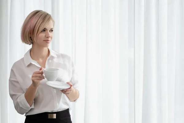 Menina loira bonita nova com cabelo curto em uma camisa branca e óculos está sentado no sofá em brilhante no escritório contra a janela. Segura uma taça branca e um pires. Beber café ou chá em um — Fotografia de Stock