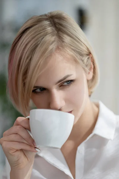 Menina loira bonita nova com cabelo curto em uma camisa branca e óculos está sentado no sofá em brilhante no escritório contra a janela. Segura uma taça branca e um pires. Beber café ou chá em um — Fotografia de Stock