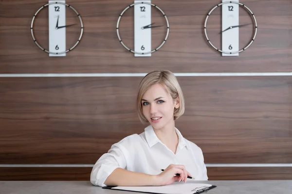 Jonge mooie blonde meid met kort haar in een wit overhemd. De receptioniste bij de receptie van het Hotel. — Stockfoto