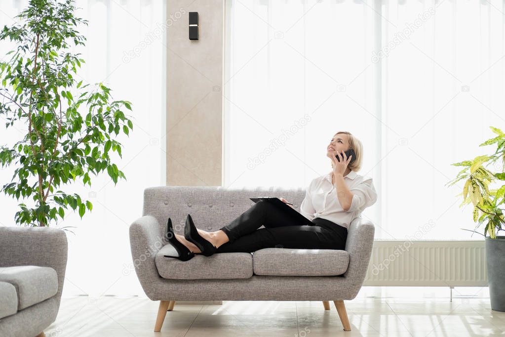 Young beautiful blonde girl with short hair in a white shirt is sitting on the sofa in bright in the office against the window. Holds a notebook and smartphone. Talking on the phone.