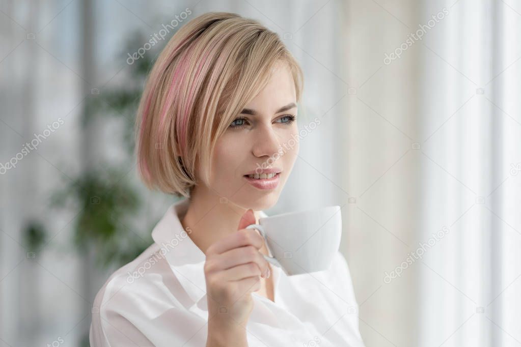 Young beautiful blonde girl with short hair in a white shirt and glasses is sitting on the sofa in bright in the office against the window. Holds a white cup and saucer. Drinking coffee or tea in a