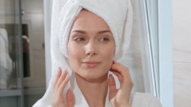 Kafasından beyaz havlu ile çekici orta yaşlı sarışın kadın ve ayna banyoda ayakta bornoz. O cilde dokunur ve gülümsüyor.