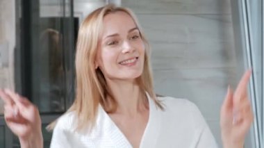 Ayna tarafından banyoda ayakta beyaz bornoz çekici orta yaşlı sarışın kadın. Komik dans ve gülümseyen.