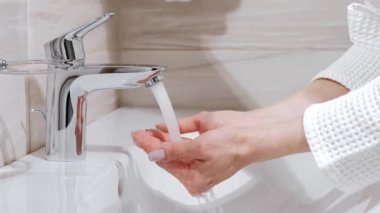 Beyaz cüppe giymiş bir kadın, ellerini banyo lavaboda yıkıyor. Musluk su akar.