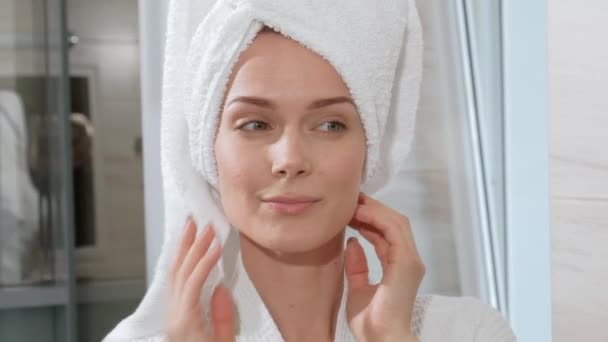 Attractive middle-aged blond woman with white towel on her head and in bathrobe standing in the bathroom by the mirror. She touches skin and smiles. — Stock Video