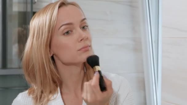 Atractiva mujer rubia de mediana edad en albornoz blanco parada en el baño junto al espejo. Aplica maquillaje . — Vídeos de Stock