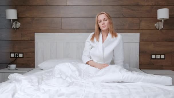 Young attractive middle-aged blonde woman in a white coat wakes up in the morning in bed. — Stock Video