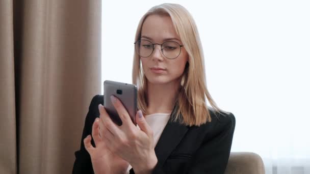 Joven mujer rubia atractiva de mediana edad en un traje de negocios negro y anteojos en una habitación de hotel. Escribe mensajes en el teléfono inteligente, funciona . — Vídeos de Stock