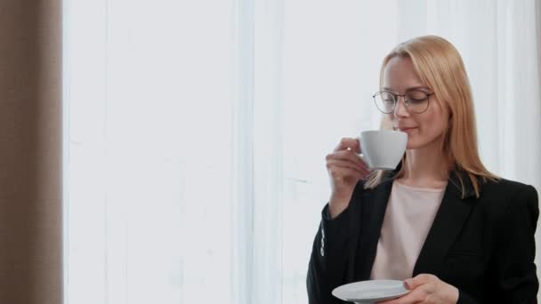 Young attractive middle-aged blonde woman in a black business suit and glasses in a hotel room. Stands by the window, drinks coffee from a white cup, looks out of the window and smiles. — Stock Video