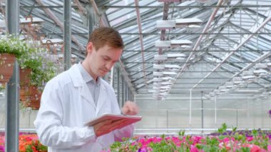 Beyaz bir ceket, bir bilim adamı biyolog veya agronomist bir genç adam inceliyor ve sera içinde çiçek ve yeşil bitkiler analiz.