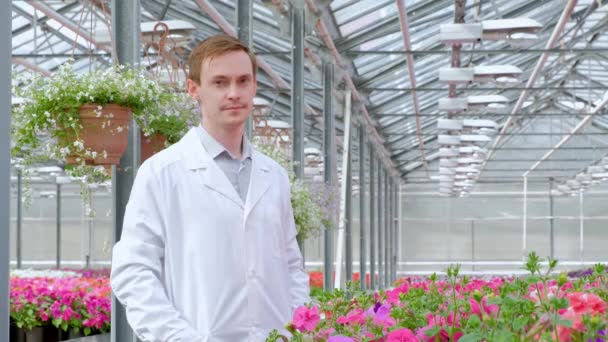 Un joven con una bata blanca, un biólogo científico o agrónomo examina y analiza las flores y plantas verdes en el invernadero . — Vídeos de Stock