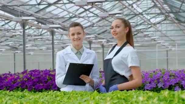 Δύο γυναίκες με άσπρα παλτά και μαύρες ποδιές. Επιστήμονες, βιολόγοι ή Γεωπόνοι εξετάζουν και αναλύουν λουλούδια και πράσινα φυτά στο θερμοκήπιο. Εγγραφή δεδομένων στο tablet. — Αρχείο Βίντεο