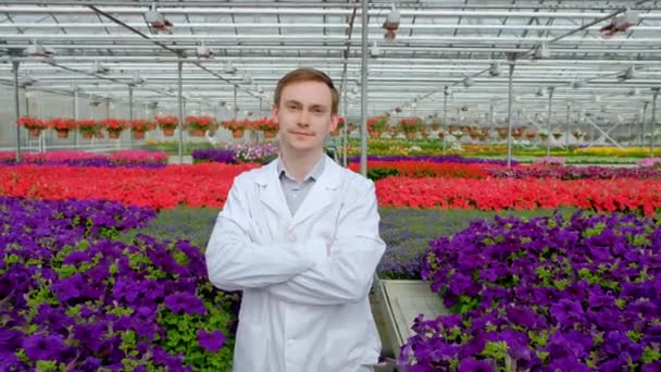 Junge männliche Wissenschaftler oder Agronomen in weißem Kittel vor dem Hintergrund der Beete mit Blumen im Gewächshaus. Blick in die Kamera. — Stockvideo