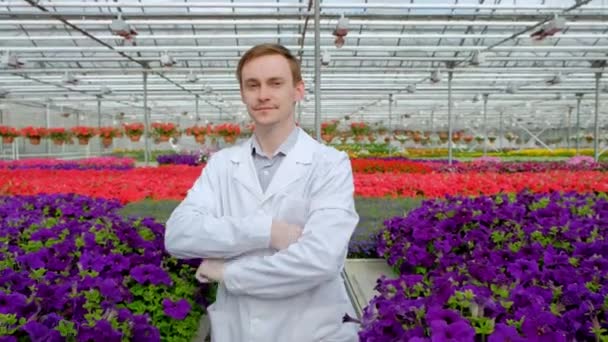 Mladý mužský vědec nebo agronomista v bílém plášti proti pozadí postelí s květinami ve skleníku. Dívám se na fotoaparát. — Stock video