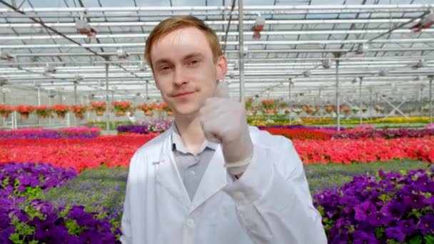 Junge männliche Wissenschaftler oder Agronomen in weißem Kittel vor dem Hintergrund der Beete mit Blumen im Gewächshaus. Blick in die Kamera. — Stockvideo