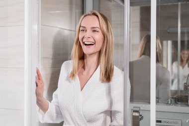 Beyaz bornozda çekici orta yaşlı sarışın kadın banyoda duş gider. Gülümsüyor ve gülüyor.