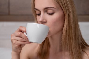 Beyaz bornozlu çekici orta yaşlı sarışın bir kadın yatak odasında bir yatağa yatıyor. Beyaz bir bardağa kahve içmek. Gülümsüyor ve gülüyor.