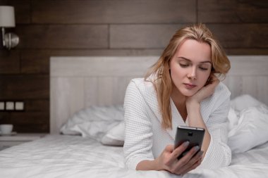 Beyaz bir bornozda çekici bir orta yaşlı sarışın kadın bir yatak odasında bir yatakta yatıyor ve bir akıllı telefon kullanarak. Gülümsüyor ve gülüyor.