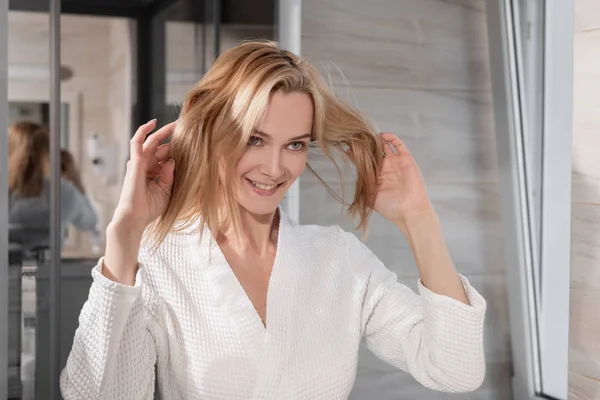 Atraktivní blondýnka středního věku v bílém plášti stojí před zrcadlem v koupelně. Dotýkala se vlasů rukama. Úsměvy a smích. — Stock fotografie