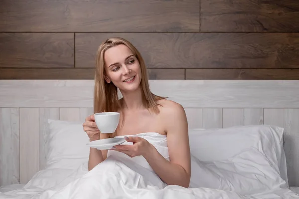 Eine attraktive blonde Frau mittleren Alters im weißen Bademantel liegt auf einem Bett in einem Schlafzimmer. Kaffee trinken aus einer weißen Tasse. lächelt und lacht. — Stockfoto