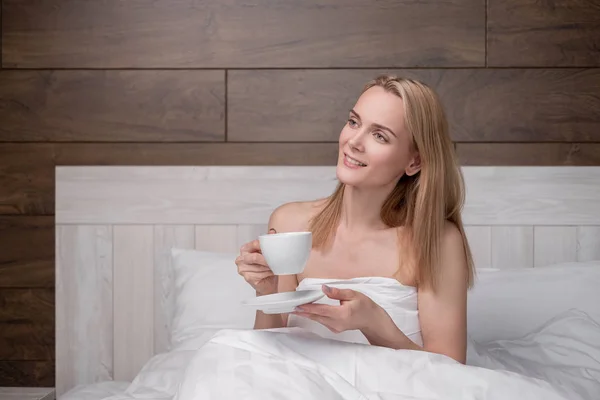 Eine attraktive blonde Frau mittleren Alters im weißen Bademantel liegt auf einem Bett in einem Schlafzimmer. Kaffee trinken aus einer weißen Tasse. lächelt und lacht. — Stockfoto