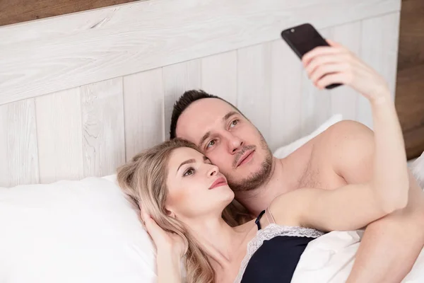 A married couple, a man and a woman, lie in bed, hug, embrace and take a selfie. — Stock Photo, Image