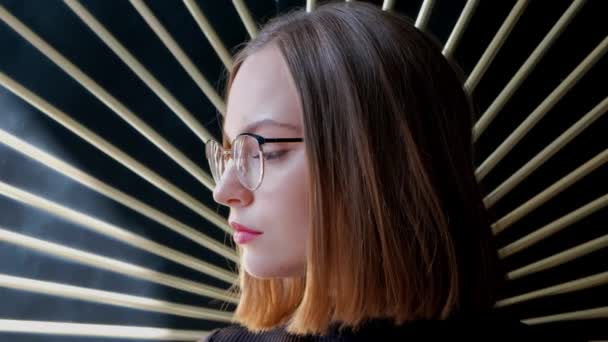 Atractiva mujer joven pelirroja con un corte de pelo corto en una plaza, con gafas y un cuello alto marrón contra una pared con una decoración de rayos dorados . — Vídeos de Stock
