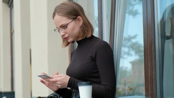 Atraktivní zrzavá mladá žena s krátkým účesem, čtyřstranná, s brýlemi a hnědou turtleneckem, sedí na lavičce u stolu v pouliční kavárně. — Stock video