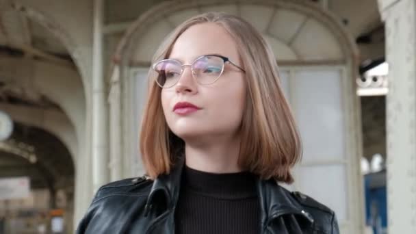 Attraktiv rödhårig ung kvinna med kort frisyr, bär glasögon och svart skinnjacka står vid vackra historiska Vitebsk järnvägsstation i St Petersburg. — Stockvideo