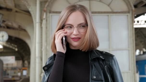 Jolie jeune femme aux cheveux roux avec une coupe courte, portant des lunettes et une veste en cuir noir se tient à la belle gare historique de Vitebsk à Saint-Pétersbourg et attend le train . — Video