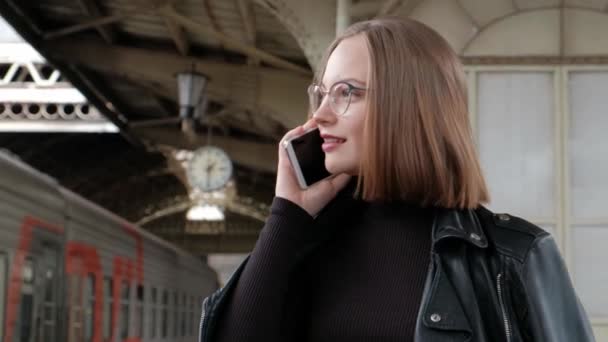 Vonzó vörös hajú fiatal nő, rövid hajvágás, szemüveg viselése és fekete bőr kabát áll a gyönyörű történelmi Vitebsk vasútállomás Szentpéterváron, és várja a vonat. — Stock videók