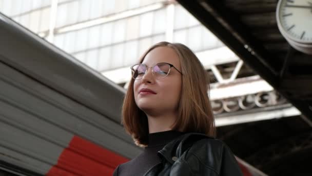 Jolie jeune femme aux cheveux roux avec une coupe courte, portant des lunettes et une veste en cuir noir se tient à la belle gare historique de Vitebsk à Saint-Pétersbourg et attend le train . — Video