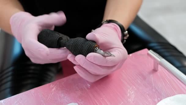 O mestre da tatuagem insere uma agulha na máquina de tatuagem. Luvas de segurança rosa em um fundo vermelho . — Vídeo de Stock