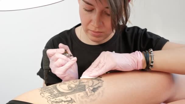 Tetovaný Mistr atraktivní žena s tetováním vytetovaní klientky na boku dívky. Černý tetování a inkoust. Obrázek rozzlobené jeptišky. Růžové a bílé studiové pozadí zdi. — Stock video
