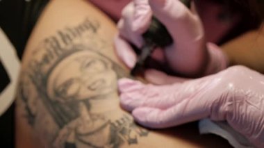 Kalça üzerinde kız dreadlocks dövmeler istemci ile Tattoo Master çekici kadın. Siyah dövme makine ve mürekkep. Öfkeli bir rahibenin resmi. Pembe ve beyaz stüdyo duvar arka plan.