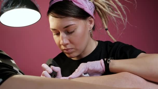 Tattoo Master attraktiv kvinna med dreadlocks tatueringar klient till flickan på höften. Svart tatuering maskin och bläck. Bild på en arg nunna. Rosa och vit Studio vägg bakgrund. — Stockvideo