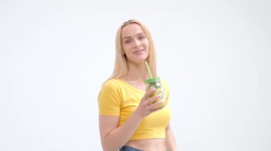 Beyaz bir arka planda poz ve bir saman ile bir cam kavanoz limonata taze bir kokteyl içme sarı bir T-shirt ve kot çekici genç sarışın kadın.