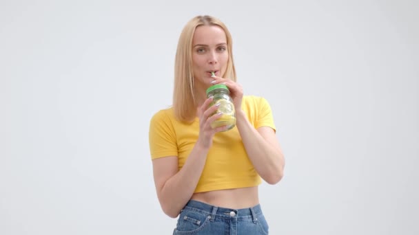 Atractiva joven rubia con una camiseta amarilla y jeans posando sobre un fondo blanco y bebiendo un cóctel fresco de limonada de un frasco de vidrio con una paja . — Vídeo de stock