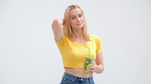 Aantrekkelijke jonge blonde vrouw in een geel T-shirt en jeans poseren op een witte achtergrond en het drinken van een frisse cocktail van limonade uit een glazen pot met een rietje. — Stockvideo