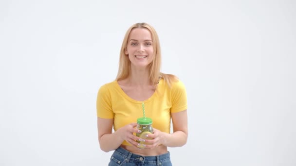 Jolie jeune femme blonde dans un T-shirt jaune et un jean posant sur un fond blanc et buvant un cocktail frais de limonade dans un pot en verre avec une paille . — Video