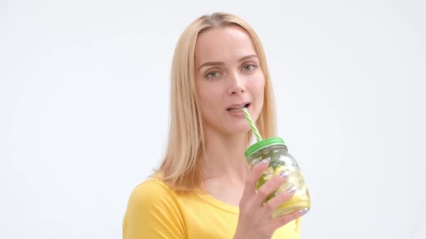 Attractive young blond woman in a yellow T-shirt and jeans posing on a white background and drinking a fresh cocktail of lemonade from a glass jar with a straw. — Stock Video