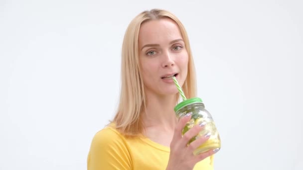 Atraktivní Mladá blondýnka ve žlutém tričku a džínách, které se na bílém pozadí vydávala a popíjejí čerstvý koktejl z limonády ze skleněné sklenice s slámou.