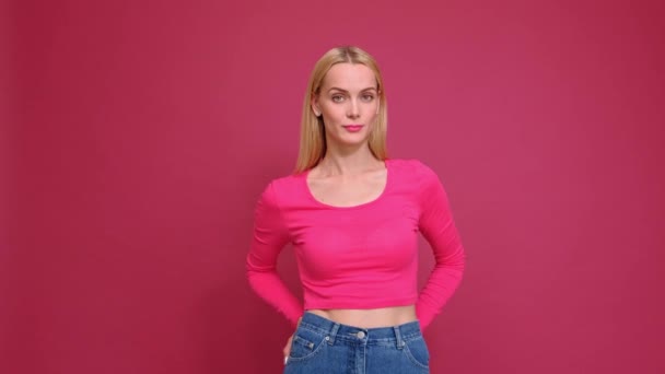 Приваблива молода жінка блондинка в червоній футболці і джинсах позує на рожевому тлі. Показує різні емоції, сюрприз, радість, смуток . — стокове відео