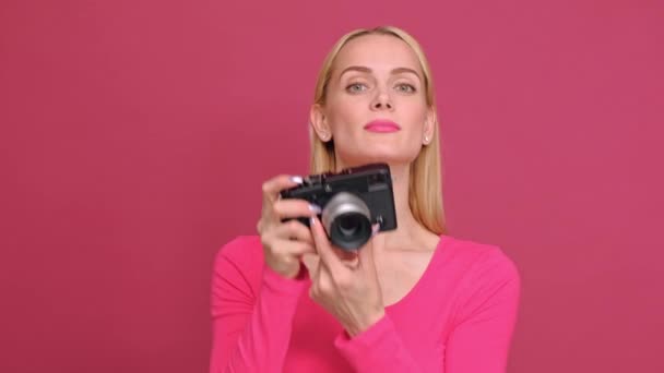 Attraktive junge blonde Frau in rotem T-Shirt und Jeans, die auf rosa Hintergrund posiert. Fotos auf einer spiegellosen Kamera im Retro-Stil und lächelnd. — Stockvideo