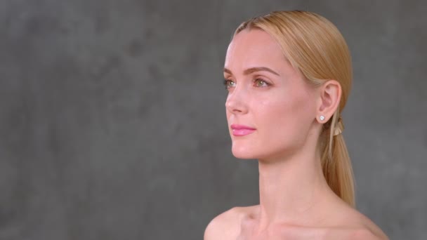 Attraktive junge blonde Frau mit nackten Schultern posiert vor grauem Hintergrund im Studio. — Stockvideo