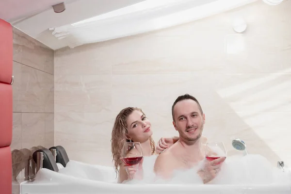 An attractive couple, a man and a woman, a husband and wife, lie in a bubble bath and drink red wine. — Stock Photo, Image