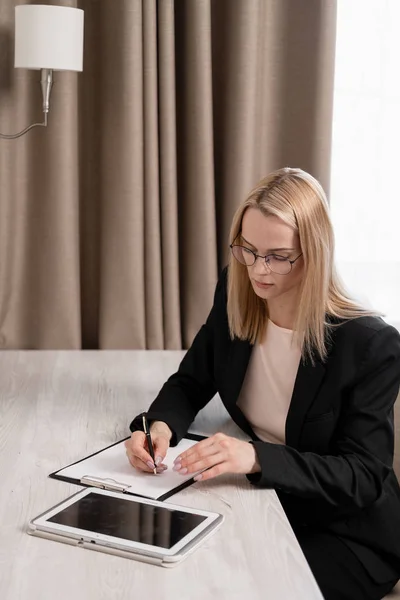Aantrekkelijke blonde vrouw in glazen en een Business Suit werken op een Tablet, praten over de telefoon en het maken van aantekeningen in een notebook. Hotel kamer, tafel bij het raam. — Stockfoto